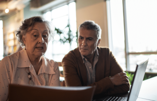 An older man and women look at a computer screen while sitting at a table together.