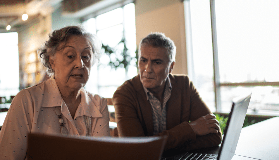 An older man and women look at a computer screen while sitting at a table together.