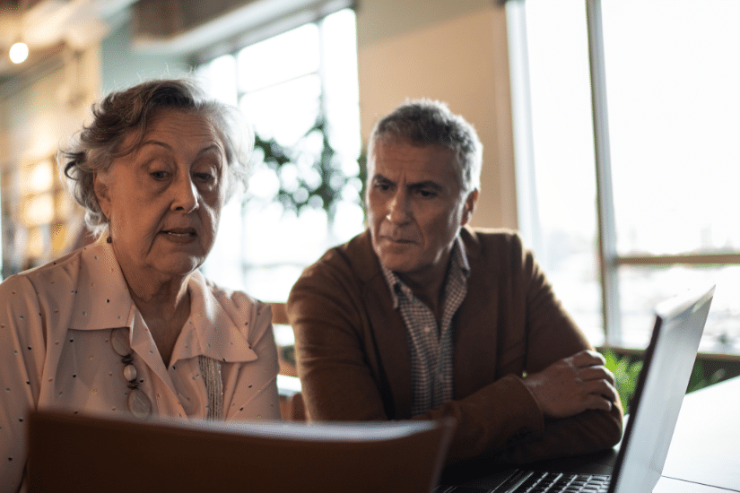 An older man and women look at a computer screen while sitting at a table together.