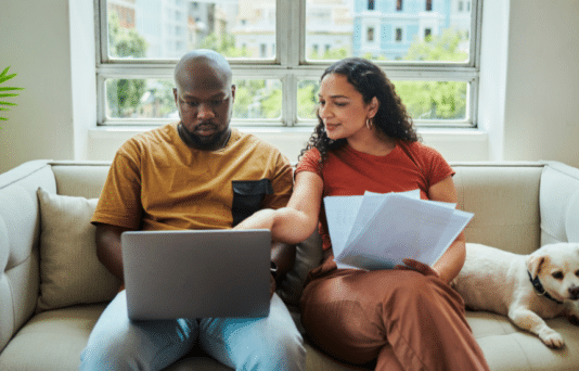 A young couple working on their budget and finances on a sofa from home with their dog using a laptop computer