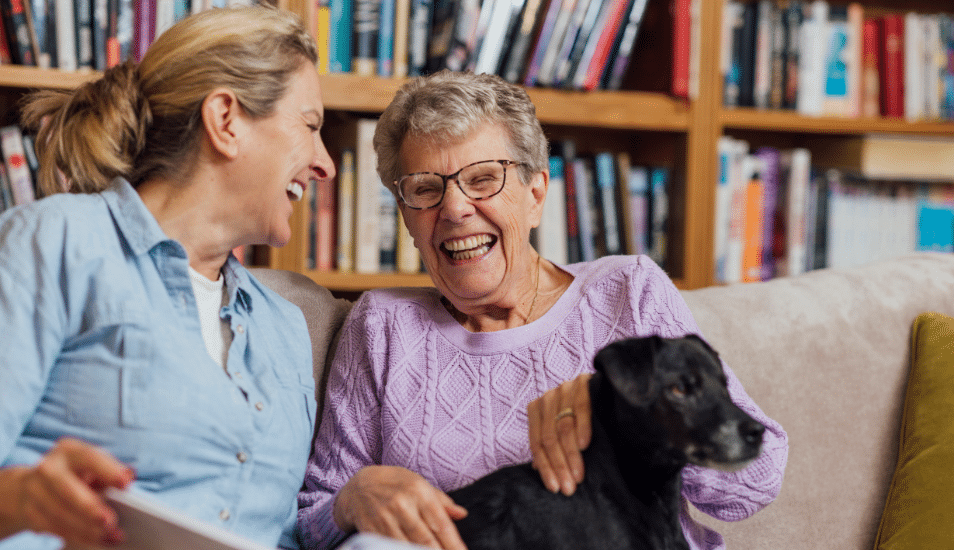 A woman sitting with her mother and pet dog on a sofa at home. They are looking at a book together and laughing.