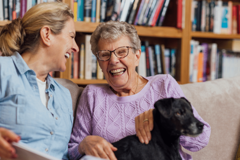 A woman sitting with her mother and pet dog on a sofa at home. They are looking at a book together and laughing.