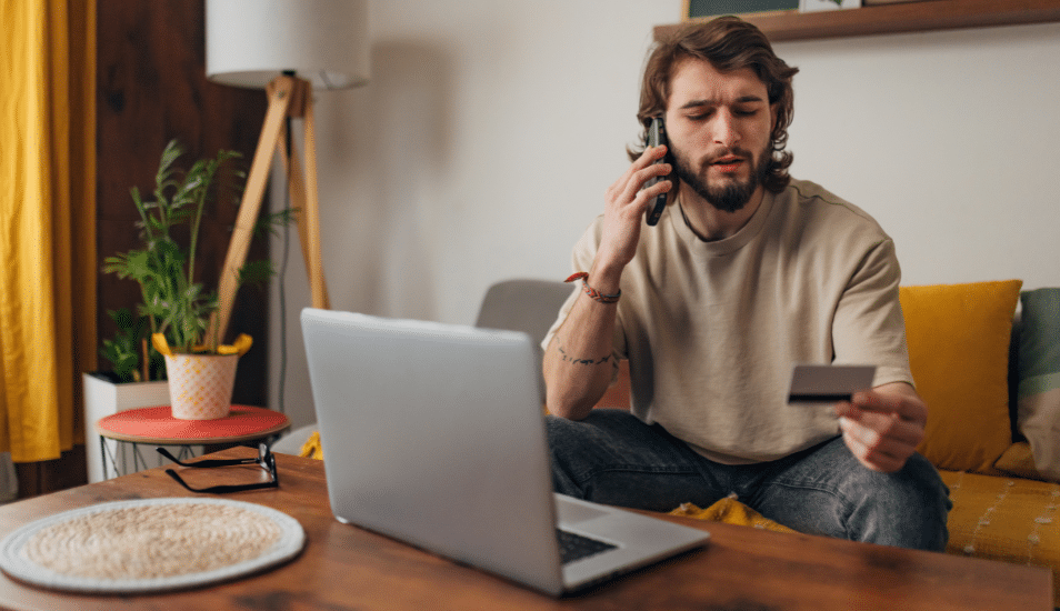 Man makes a phone call while trying to make an online purchase using a credit card and a laptop.