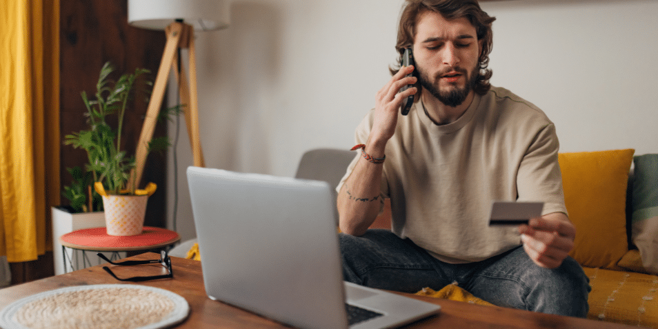 Man makes a phone call while trying to make an online purchase using a credit card and a laptop.