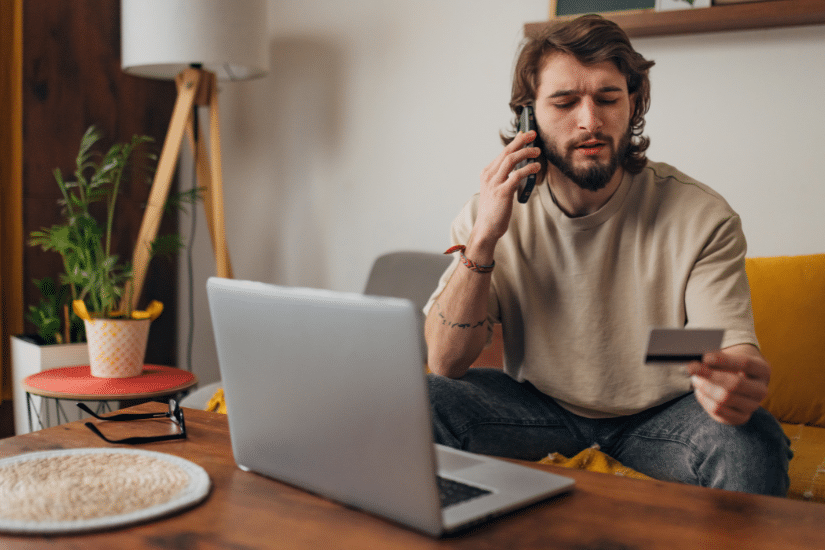 Man makes a phone call while trying to make an online purchase using a credit card and a laptop.