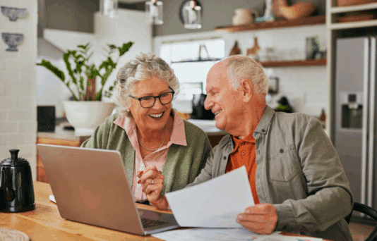 older couple sitting in the kitchen looking at documents and a computer.