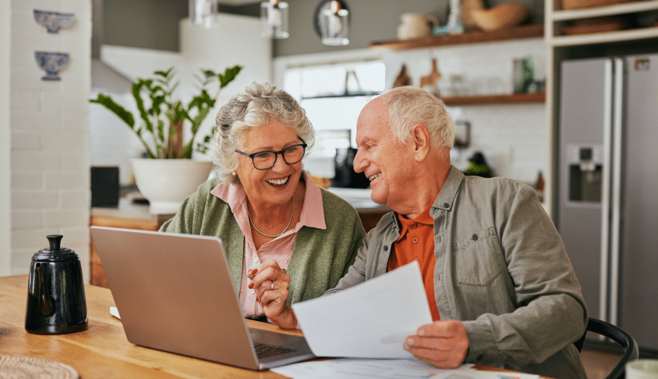 older couple sitting in the kitchen looking at documents and a computer.