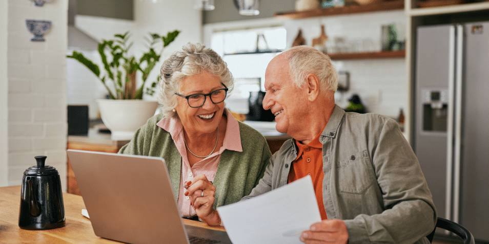 older couple sitting in the kitchen looking at documents and a computer.