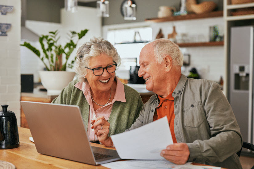 older couple sitting in the kitchen looking at documents and a computer.