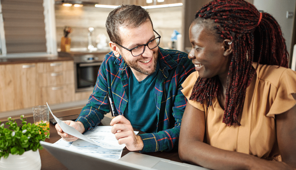 Diverse couple sitting at the kitchen table going over documents and smiling at each other.
