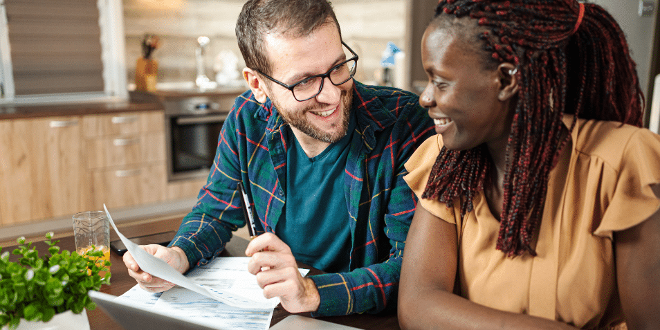 Diverse couple sitting at the kitchen table going over documents and smiling at each other.