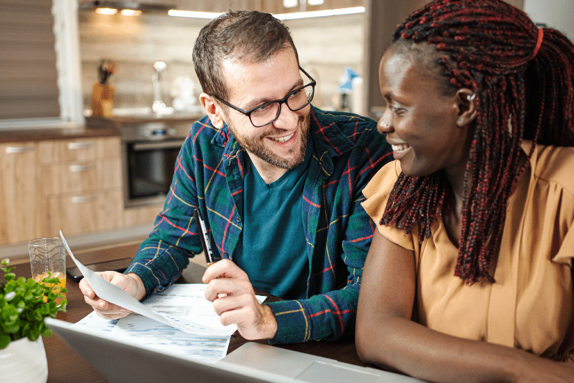 Diverse couple sitting at the kitchen table going over documents and smiling at each other.