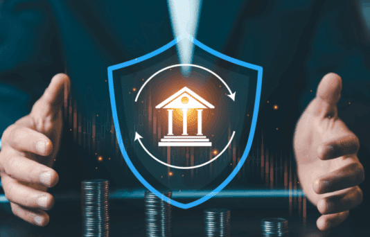 Businessman protecting bank icon inside blue shield with stacks of coins, financial insurance and capital protection concept against market volatility.