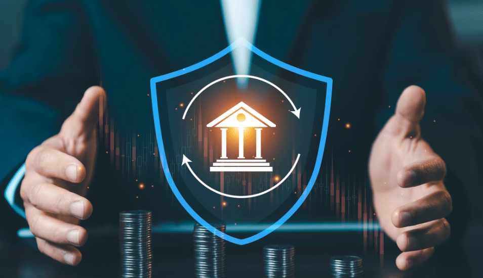 Businessman protecting bank icon inside blue shield with stacks of coins, financial insurance and capital protection concept against market volatility.