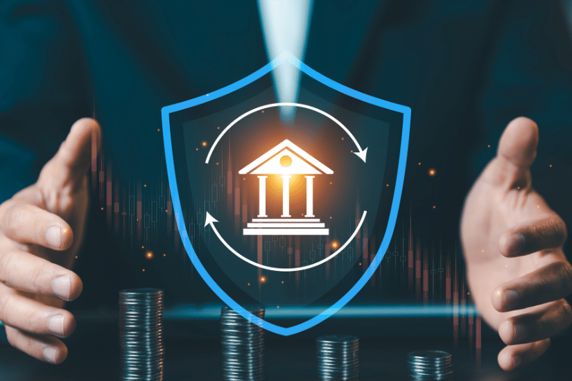 Businessman protecting bank icon inside blue shield with stacks of coins, financial insurance and capital protection concept against market volatility.